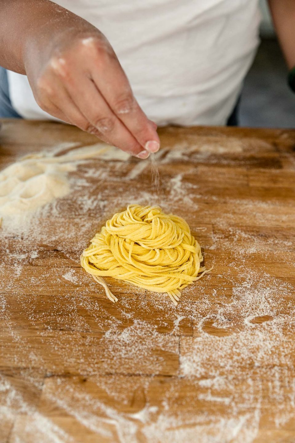 A bundle of fresh pasta cut into spaghetti rests atop a butcher block countertop that is dusted with semolina flour. A woman's hand dusts additional semolina flour over the bundle.