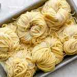 An overhead shot of six bundles of fresh pasta cut into spaghetti noodles are arranged on a small aluminum baking sheet and dusted with semolina flour. The baking sheet sits atop a white textured surface. A light gray linen napkin is positioned alongside the baking sheet.