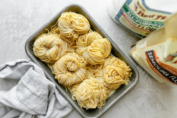 An overhead shot of six bundles of fresh pasta cut into spaghetti noodles are arranged on a small aluminum baking sheet and dusted with semolina flour. The baking sheet sits atop a white textured surface. One bag of Bob's Red Mill Organic Unbleached White All-Purpose flour, Bob's Red Mill Semolina flour, & a light gray linen napkin surround the baking sheet.