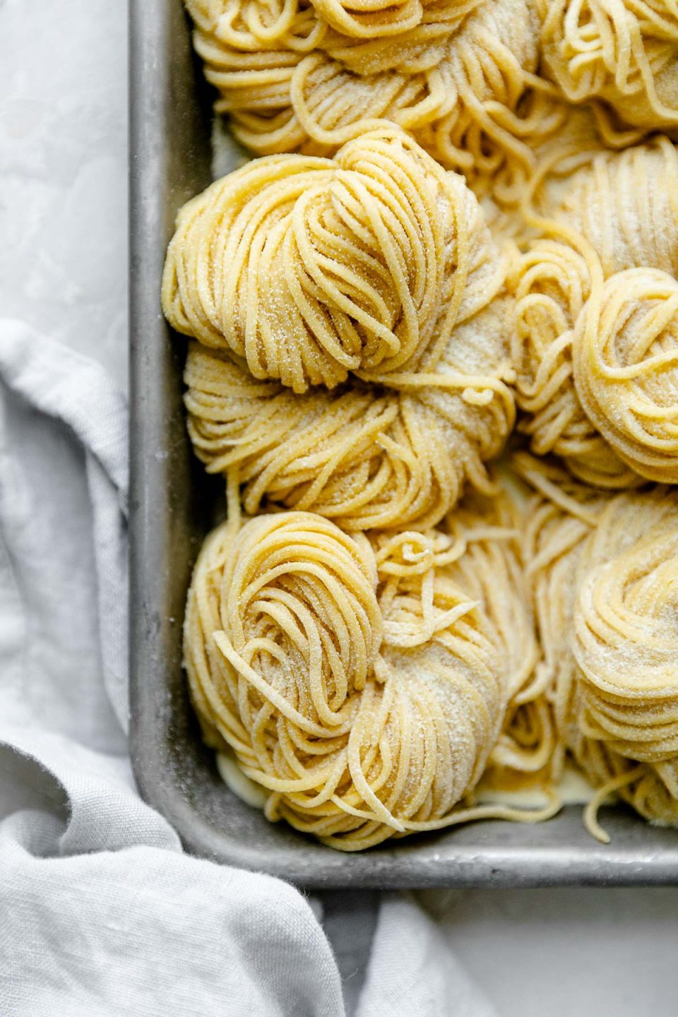A close up of bundles of fresh pasta cut into spaghetti noodles are arranged on a small aluminum baking sheet and dusted with semolina flour. The baking sheet rests atop a white textured surface and a light blue linen napkin rests alongside the baking sheet.