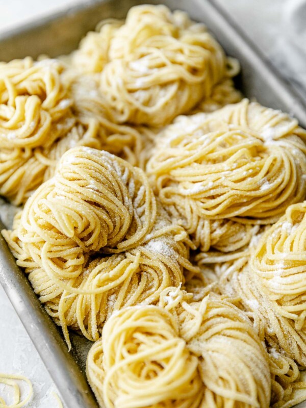 A side angle shot of six bundles of fresh pasta cut into spaghetti noodles are arranged on a small aluminum baking sheet and dusted with semolina flour. The baking sheet sits atop a white textured surface. A light blue napkin & a few stray noodles surround the baking sheet.