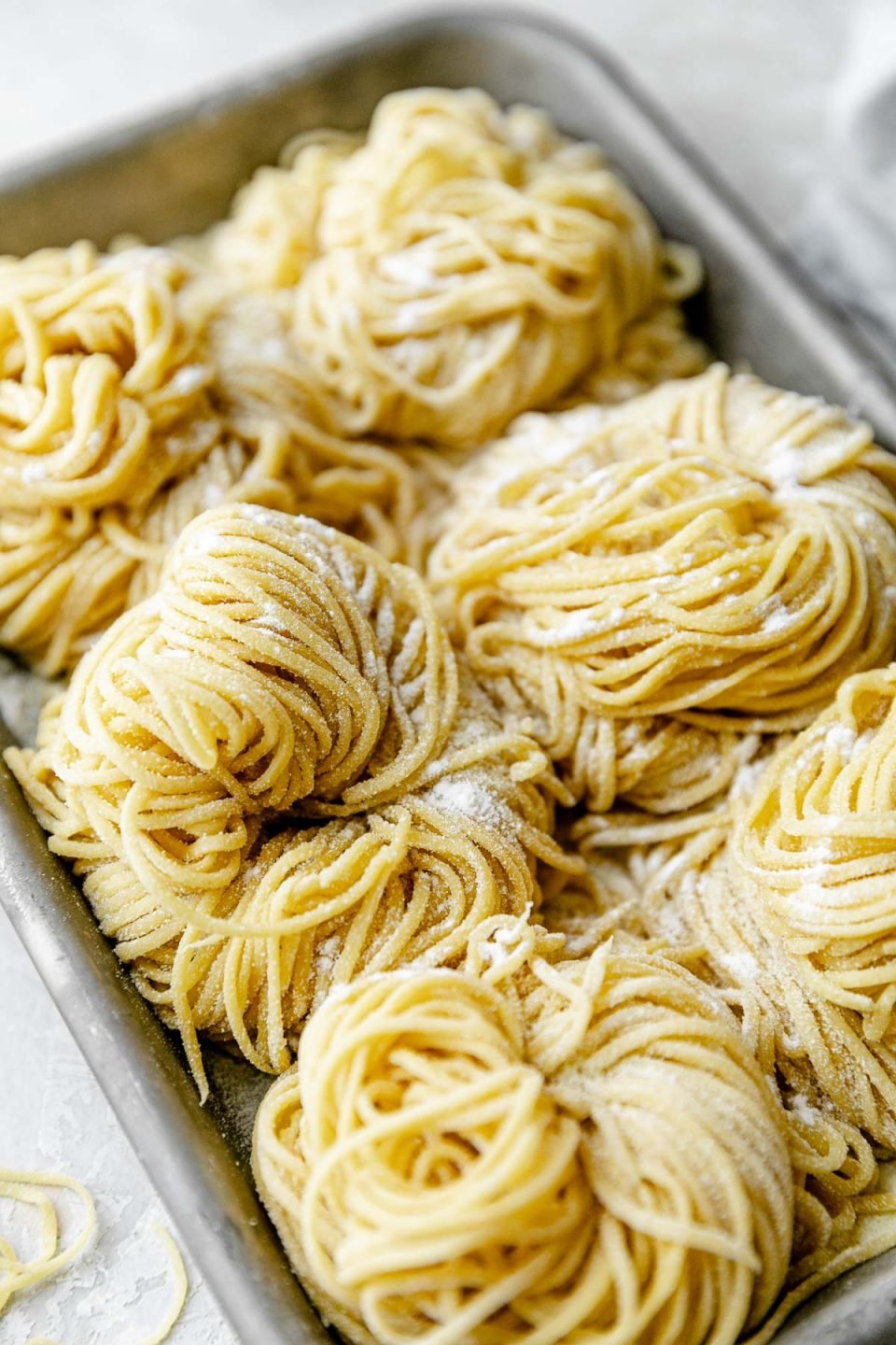 A side angle shot of six bundles of fresh pasta cut into spaghetti noodles are arranged on a small aluminum baking sheet and dusted with semolina flour. The baking sheet sits atop a white textured surface. A light blue napkin & a few stray noodles surround the baking sheet.