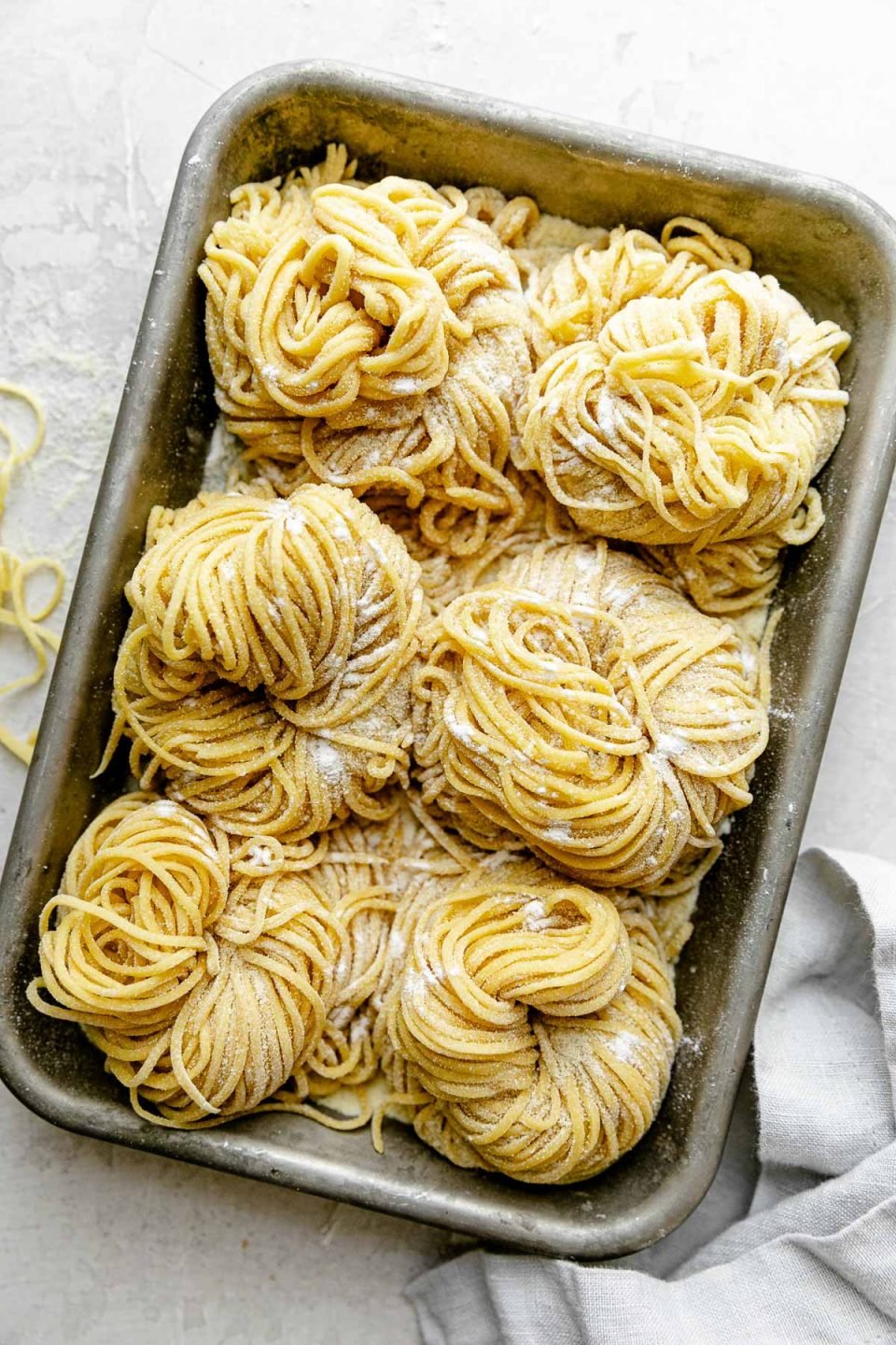 Six bundles of fresh pasta cut into spaghetti noodles are arranged on a small aluminum baking sheet and dusted with semolina flour. The baking sheet sits atop a white textured surface. A light blue napkin & a few stray noodles surround the baking sheet.