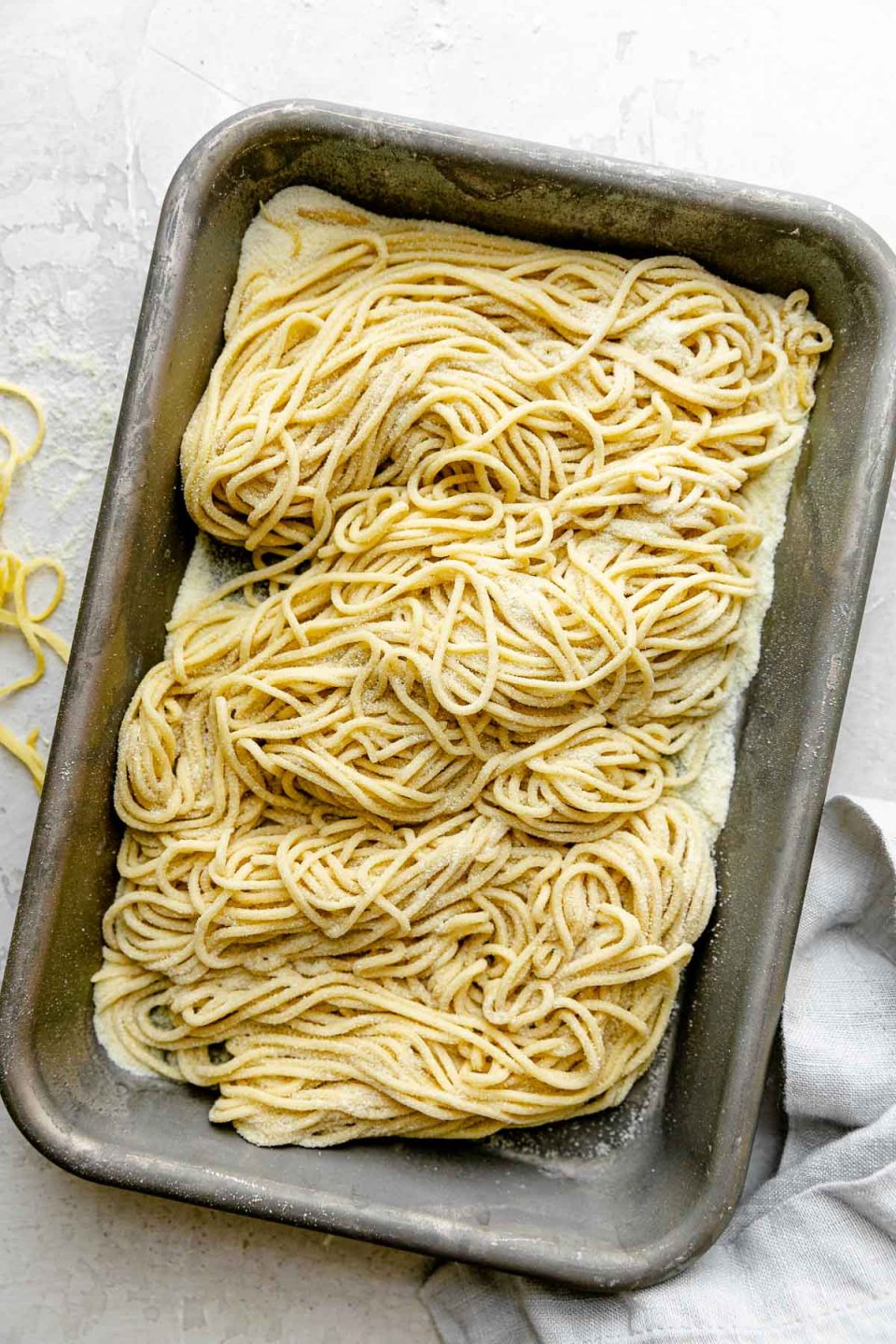 Fresh pasta cut into spaghetti noodles are arranged on a small aluminum baking sheet and dusted with semolina flour. The baking sheet sits atop a white textured surface. A light blue napkin & a few stray noodles surround the baking sheet.