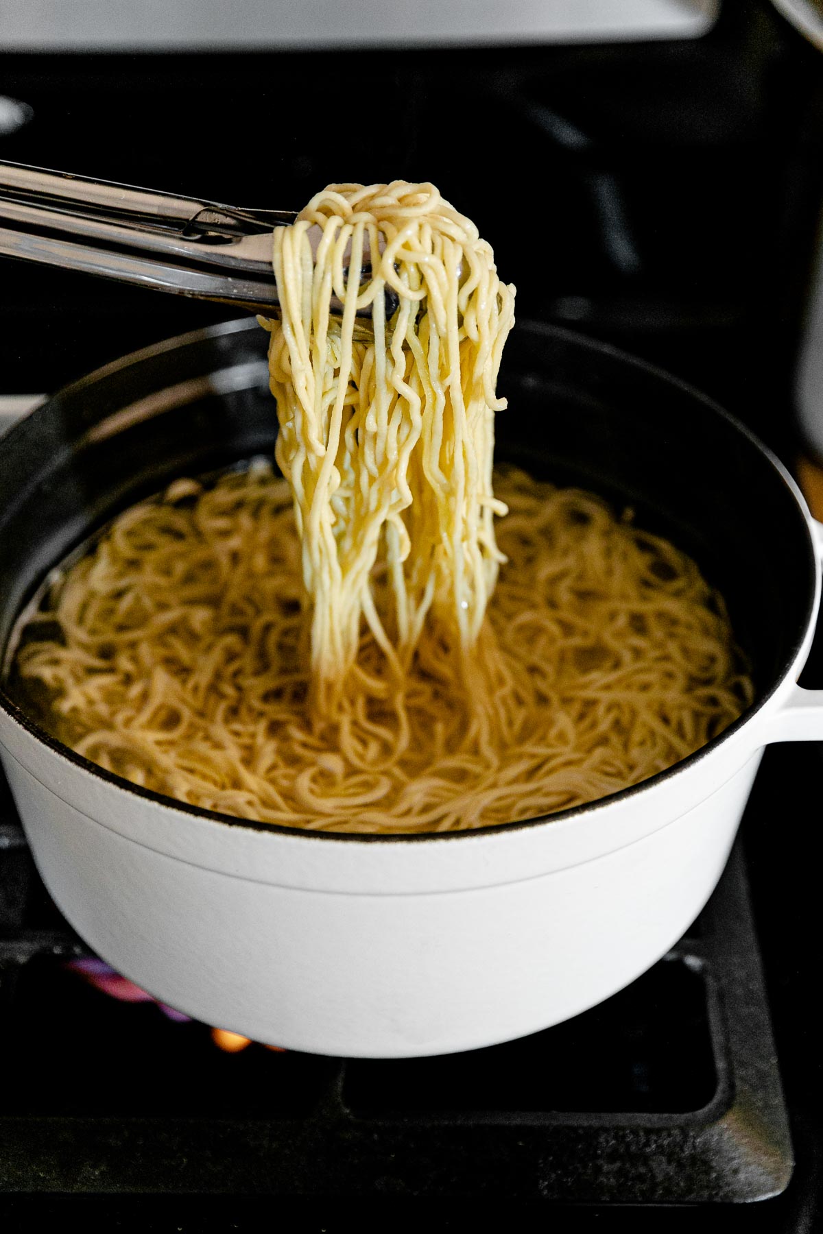 Tongs are used to pick up a bundle of fresh homemade pasta and hold it up over a white pot filled with boiling water and more fresh pasta as it cooks. The pot rests atop a gas stovetop range.