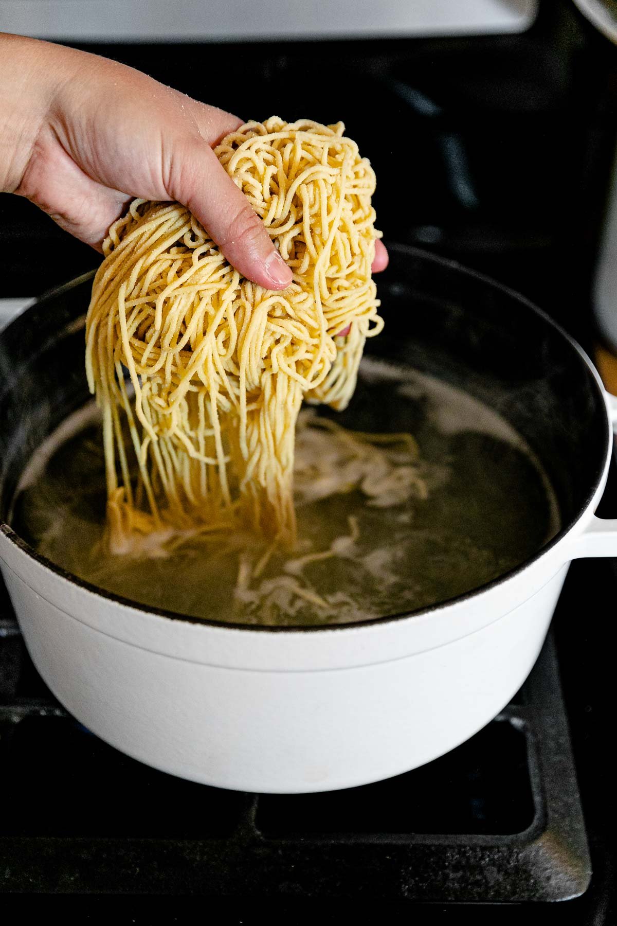 A woman's hand adds fresh homemade pasta to a white pot filled with boiling water. The pot rests atop a gas stovetop range.