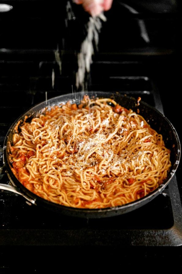 A black frying pan filled with fresh homemade pasta tossed in bolognese sauce is dusted with freshly grated parmigiano reggiano. The frying pan rests atop a gas stovetop range.
