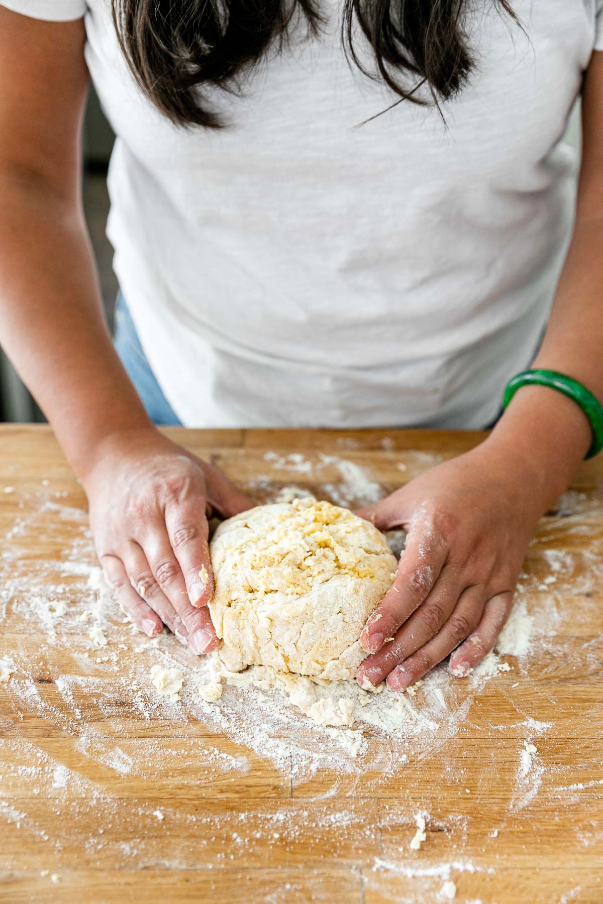 How to Make Homemade Pasta, Step 2: Mixing the Pasta Dough. Jess of Plays Well With Butter places her hands around a dough ball of fresh pasta dough. The dough ball sits atop a butcher block countertop that is dusted with flour from the process of mixing & forming the dough ball.