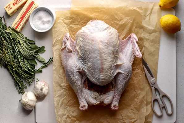 Spatchcock Turkey ingredients arranged on a light textured surface: one whole turkey, kosher salt, butter, lemon, garlic, and fresh herbs. The whole turkey rests a top two sheets of brown crumpled parchment paper that sit atop a white cutting board. A pair of beige kitchen shears rest alongside the raw turkey.