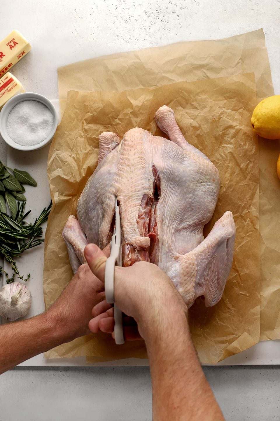 How to Spatchcock a Turkey, step 2: A man's hands hold a pair of sharp kitchen shears in one hand and hold the whole turkey's backbone with the other. He is using the kitchen shears to carefully snip alongside both sides of the turkey's backbone working to remove it completely. The turkey rests a top two sheets of brown crumpled parchment paper that sit atop a white cutting board. Two lemons, a white ramekin filled with kosher salt, two whole garlic bulbs, two sticks of butter, and fresh herbs surround the raw whole turkey.