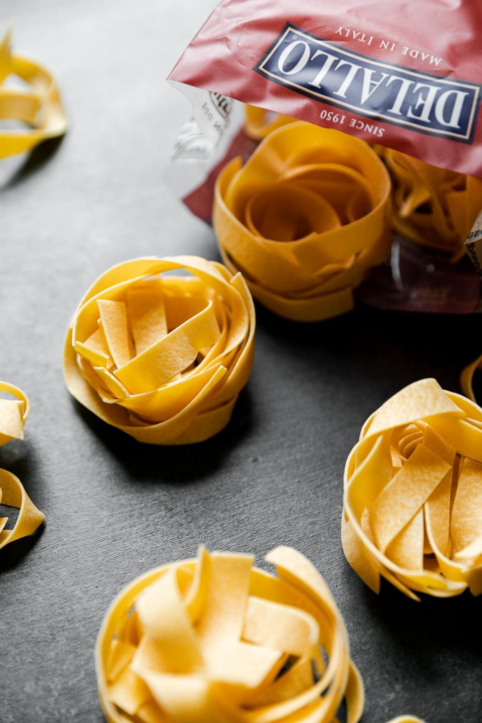 Portion-sized nests of dried DeLallo pappardelle noodles arranged on a dark textured surface resting alongside an opened bag of DeLallo Egg Pappardelle noodles.