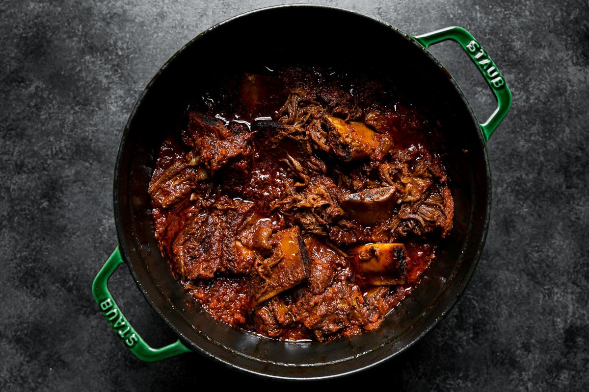 Slowly braised beef short rib ragu in a large Dutch oven atop a dark textured surface.