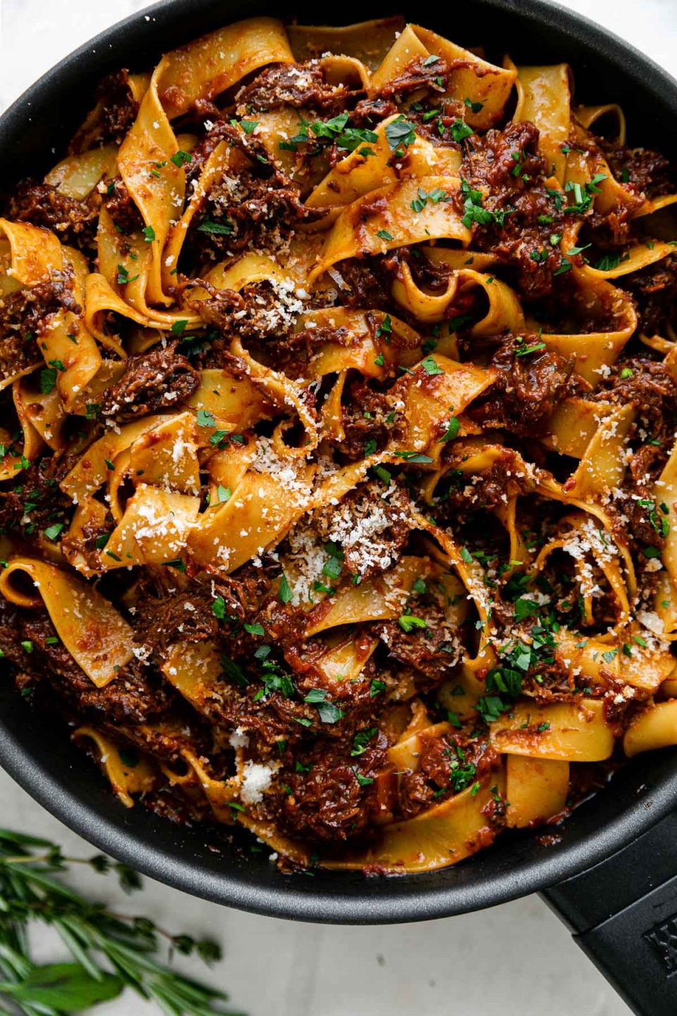 Beef short rib ragu pappardelle in a small skillet atop a light creamy textured surface. The ragu is topped with freshly grated parmesan & chopped herbs. A few sprigs of fresh herbs rest on the surface alongside the skillet.