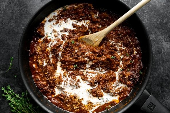 Reheating the short rib ragu with heavy cream in a small skillet. The skillet sits atop a black surface with a few sprigs of fresh herbs resting alongside of the skillet. A wooden spoon used to stir the heavy cream into the ragu sauce rests inside of the skillet.