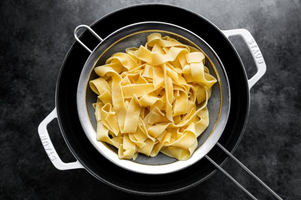 Cooked pappardelle pasta drained in a mesh strainer that rests overtop a white dutch oven. The dutch oven sits atop a black surface.