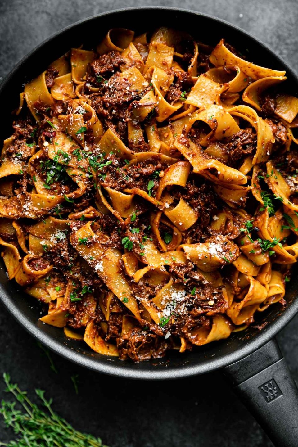 Slowly Braised Short Rib Ragu Pappardelle in a small skillet atop a black surface. The ragu has been topped with grated parmesan & fresh parsley. A few sprig of herbs rest alongside the skillet.
