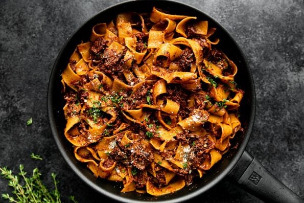 Slowly Braised Short Rib Ragu Pappardelle in a small skillet atop a black surface. The ragu has been topped with grated parmesan & fresh parsley. A few sprig of herbs rest alongside the skillet.