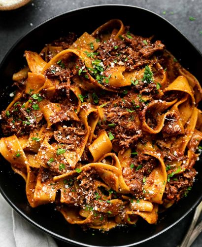 Braised Short Rib Ragu Pappardelle shown in a pasta bowl, topped with grated parmesan & fresh parsley. The bowl sits atop a black surface surrounded by a small wooden pinch bowl filled with grated parmesan, a gray linen napkin, & a set of silver silverware.