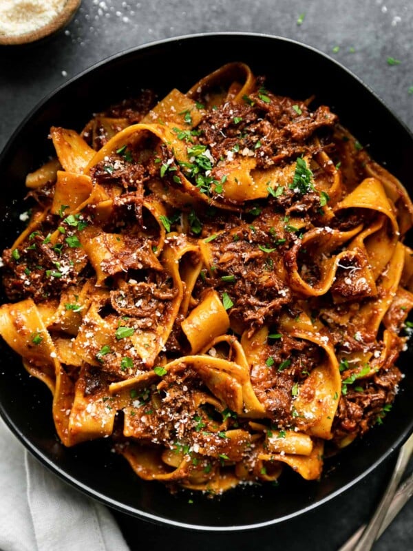 Braised Short Rib Ragu Pappardelle shown in a pasta bowl, topped with grated parmesan & fresh parsley. The bowl sits atop a black surface surrounded by a small wooden pinch bowl filled with grated parmesan, a gray linen napkin, & a set of silver silverware.