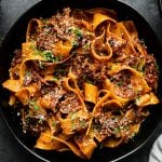 Braised Short Rib Ragu Pappardelle shown in pasta bowls, topped with grated parmesan & fresh parsley. The bowls sit atop a black surface surrounded by a clear glass filled with red wine, a gray linen napkin, & two silver forks.