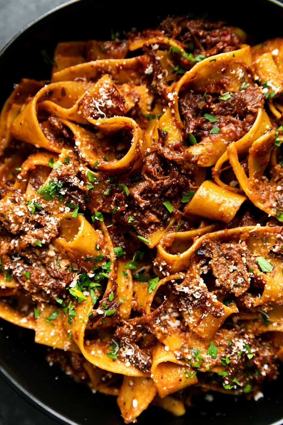 An overhead shot of Braised Short Rib Ragu Pappardelle shown in a black pasta bowl, topped with grated parmesan & fresh parsley. The bowls sit atop a black surface.