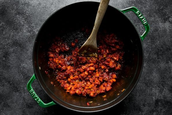 Tomato paste & aromatics mixed into browned soffritto in a large Dutch oven atop a dark textured surface. A wooden spoon rests inside the dutch oven used for stirring the soffritto.