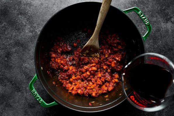 Wine is being poured into a mixture of tomato paste & aromatics mixed into browned soffritto in a large Dutch oven atop a dark textured surface. This mixture is to create a short rib ragu sauce. A wooden spoon rests inside the dutch oven used for stirring the ingredients together.