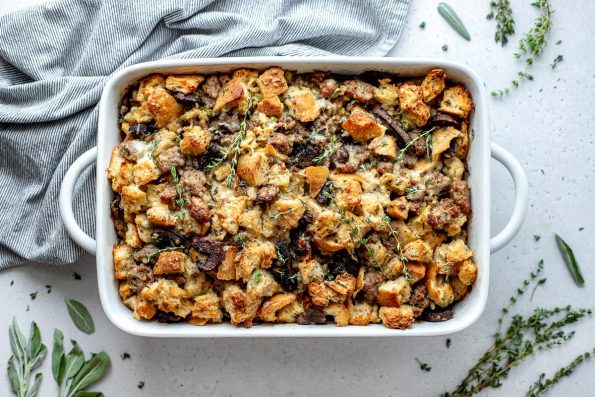 Baked sourdough stuffing with sausage and mushrooms in a white baking dish rests atop a light textured surface ​surrounded by a gray and white striped linen napkin and fresh herbs