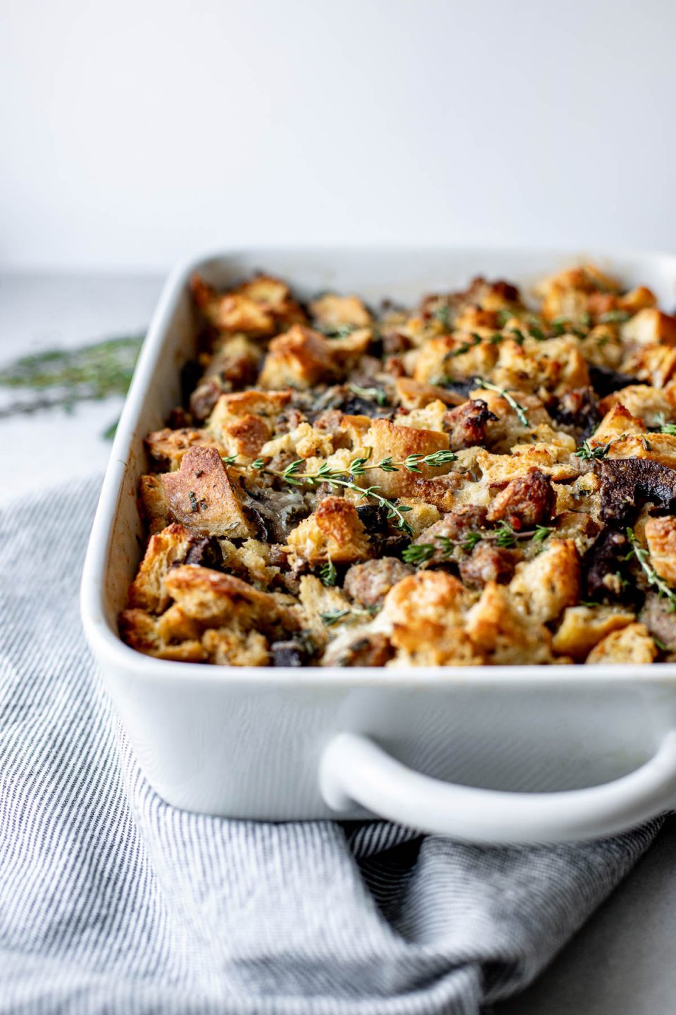 A side angle shot of baked sourdough stuffing with sausage and mushrooms in a white baking dish rests atop a light textured surface ​surrounded by a gray and white striped linen napkin and fresh herbs