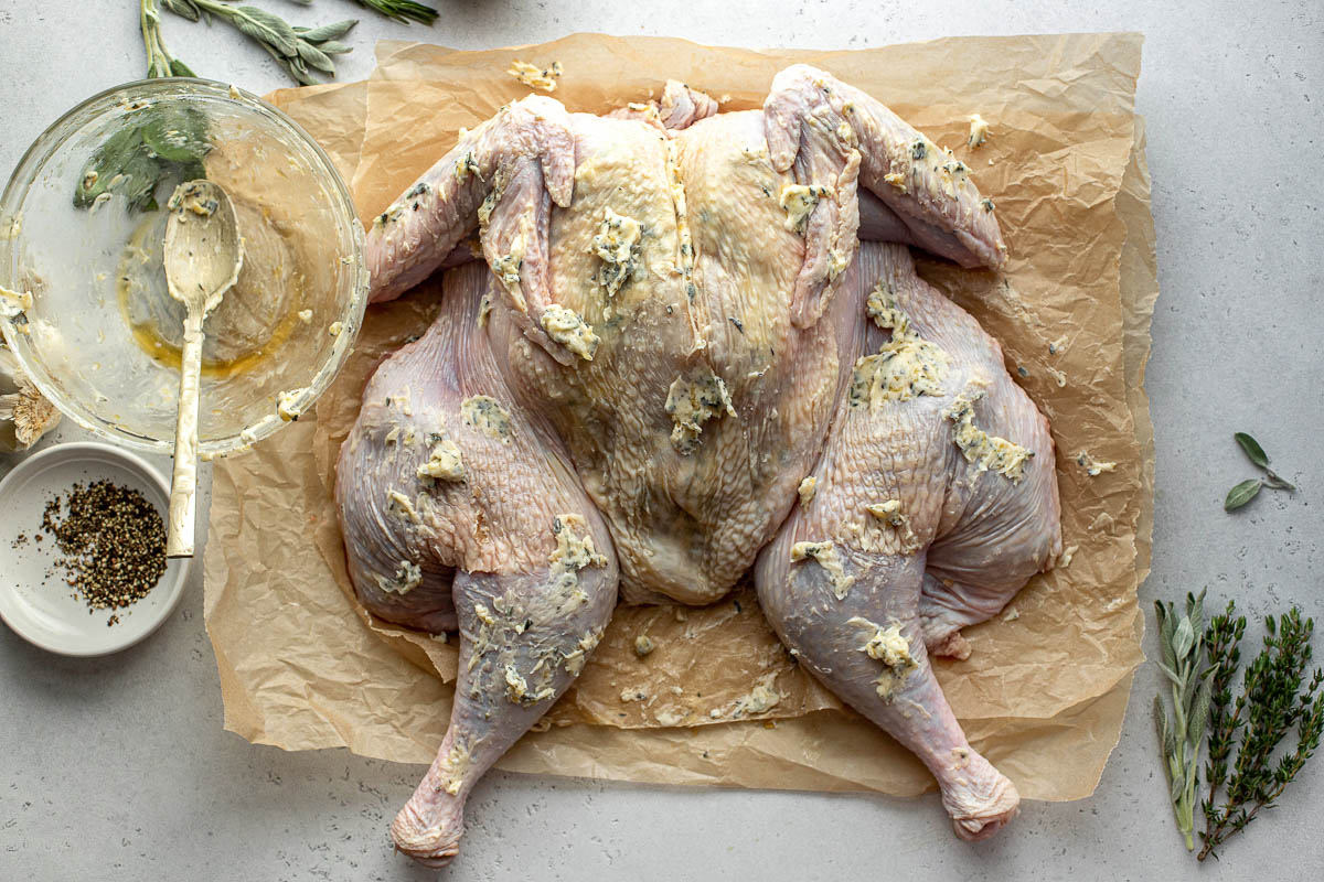 A spatchcocked turkey seasoned with maple herb butter directly on the exterior skin rests breast side up on a few sheets of brown parchment paper. A now empty bowl previously filled with prepared maple herb butter sits alongside the turkey with a gold spoon resting inside. Additionally, a few sprigs of fresh herbs, a bulb of garlic, and a small white pinch bowl filled with ground black pepper surround the turkey. All items sit atop a creamy white textured surface.