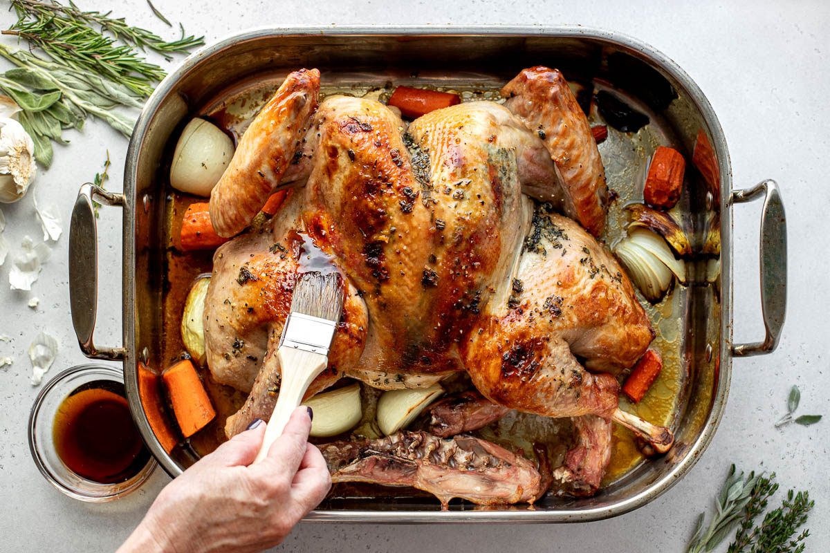 A spatchcocked turkey seasoned with maple herb butter is placed within a roasting pan surrounded by yellow onion, carrots, and the turkey neck and spine. A woman's hand holds a basting brush and is using it to baste the surface of the turkey with pure maple syrup. The roasting pan is surrounded by sprigs of fresh herbs, garlic, and a small clear glass bowl filled with pure maple syrup. All items sit atop a creamy white textured surface.