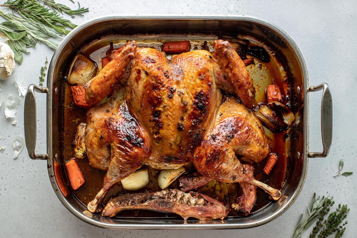 An overhead shot of a roasted Maple Glazed spatchcocked turkey seasoned with pure maple syrup and a maple herb butter placed within a roasting pan surrounded by pieces of yellow onion, carrots, and the turkey neck and spine. Turkey drippings have formed at the bottom of the roasting pan. The roasting pan is surrounded by sprigs of fresh herbs and garlic and all items sit atop a creamy white textured surface area.