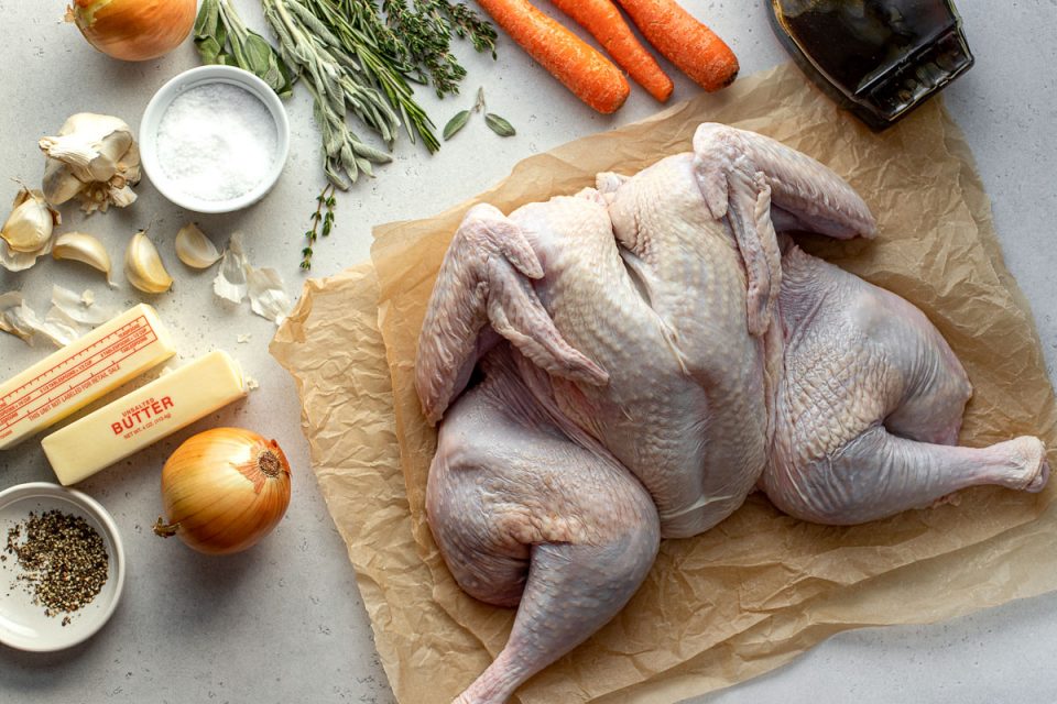 Maple Glazed Spatchcock Turkey ingredients arranged on a creamy white textured surface: a whole spatchcocked turkey lays flat on a few sheets of crumpled brown parchment paper, pure maple syrup, carrots, fresh herbs including thyme, rosemary, and sage, kosher salt, yellow onion, garlic, unsalted butter, and ground black pepper.