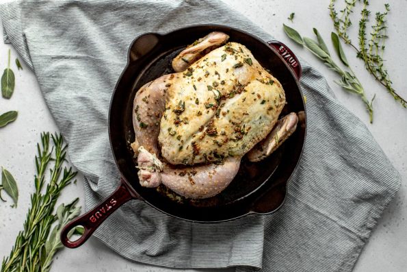 A seasoned whole chicken is placed in a large cast iron skillet for roasting. The skillet sits a top a blue and white striped linen napkin and is surrounded by piles of fresh herbs. The skillet, napkin, and herbs sit atop a lightly colored textured surface.