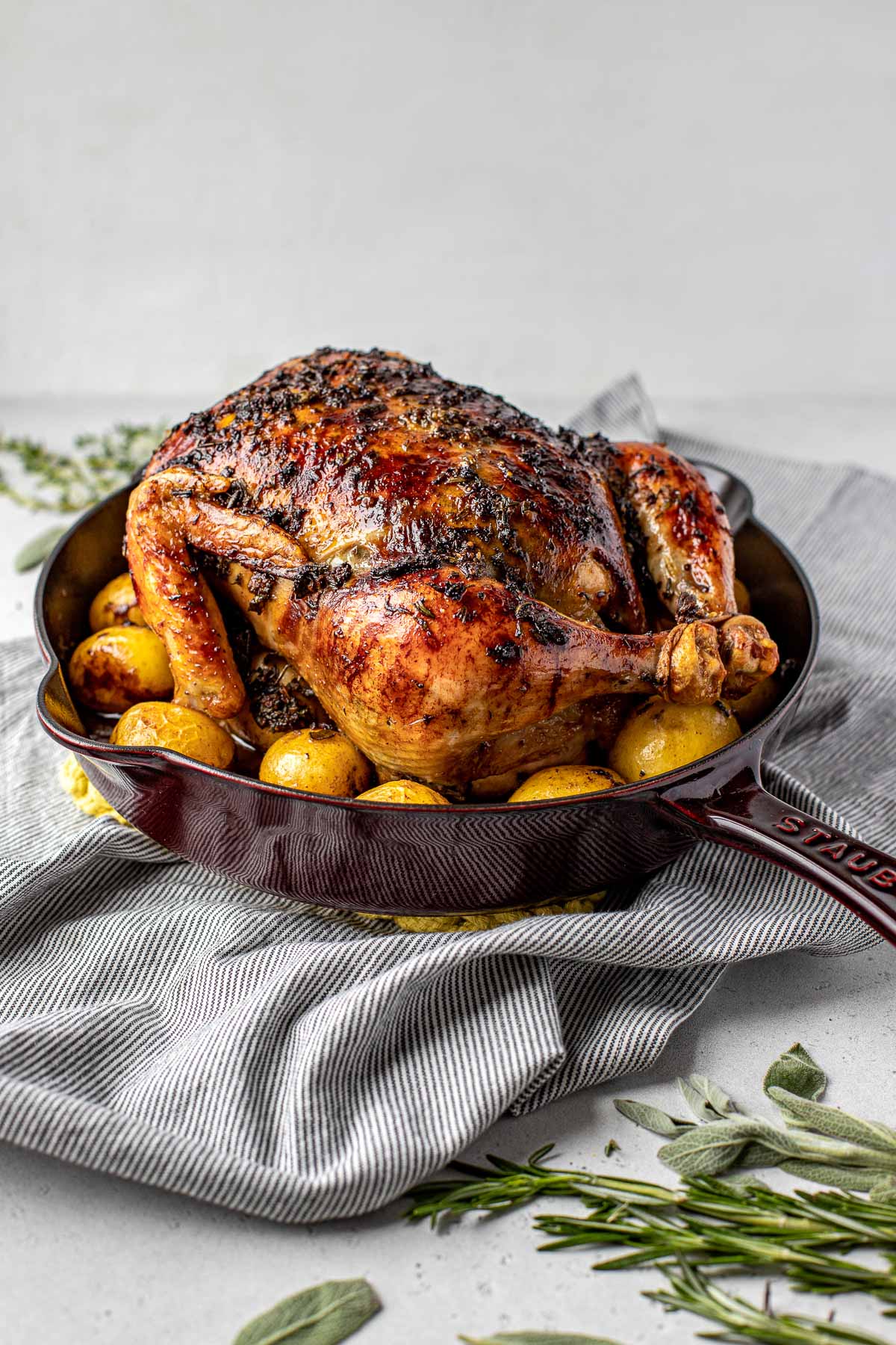 A finished roasted Thanksgiving Chicken atop a layer of roasted baby potatoes in a large cast iron skillet sits atop a blue and white striped linen napkin laid out on a lightly colored textured surface. Loose sprigs of fresh herbs surround the skillet in both the foreground and background.