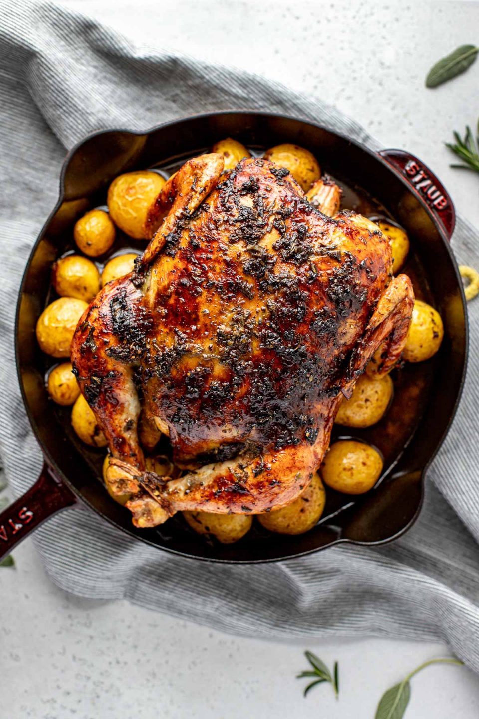 An overhead shot of a finished roasted Thanksgiving Chicken atop a layer of roasted baby potatoes in a large cast iron skillet sits atop a blue and white striped linen napkin laid out on a lightly colored textured surface. Loose sprigs of fresh herbs surround the skillet in both the foreground and background.