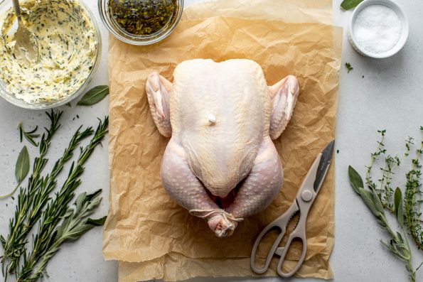Thanksgiving chicken ingredients arranged on a lightly colored textured surface: a whole chicken sits atop a couple of pieces of crumpled brown parchment paper, savory herb oil, herb butter, kosher salt, garlic, baby potatoes, and fresh herbs. A pair of kitchen shears rest atop the parchment paper alongside the whole chicken.