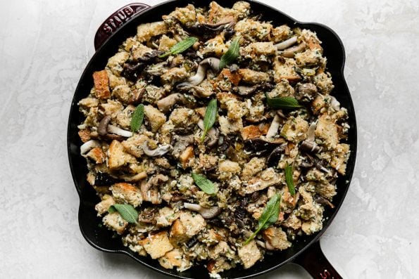 Assembled mushroom stuffing fills a Grenadine colored Staub Cast Iron Skillet that sits atop a creamy white textured surface. The stuffing is topped with fresh sage leaves prior to being baked in the oven.