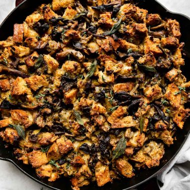 Baked wild mushroom stuffing fills a Grenadine colored Staub Cast Iron Skillet that sits atop a creamy white textured surface. A light gray linen napkin, loose sprigs of fresh herbs, and a small ceramic bowl filled with gravy sits alongside the skillet.