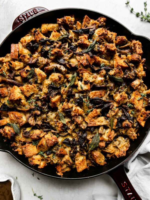 Baked wild mushroom stuffing fills a Grenadine colored Staub Cast Iron Skillet that sits atop a creamy white textured surface. A light gray linen napkin, loose sprigs of fresh herbs, and a small ceramic bowl filled with gravy sits alongside the skillet.