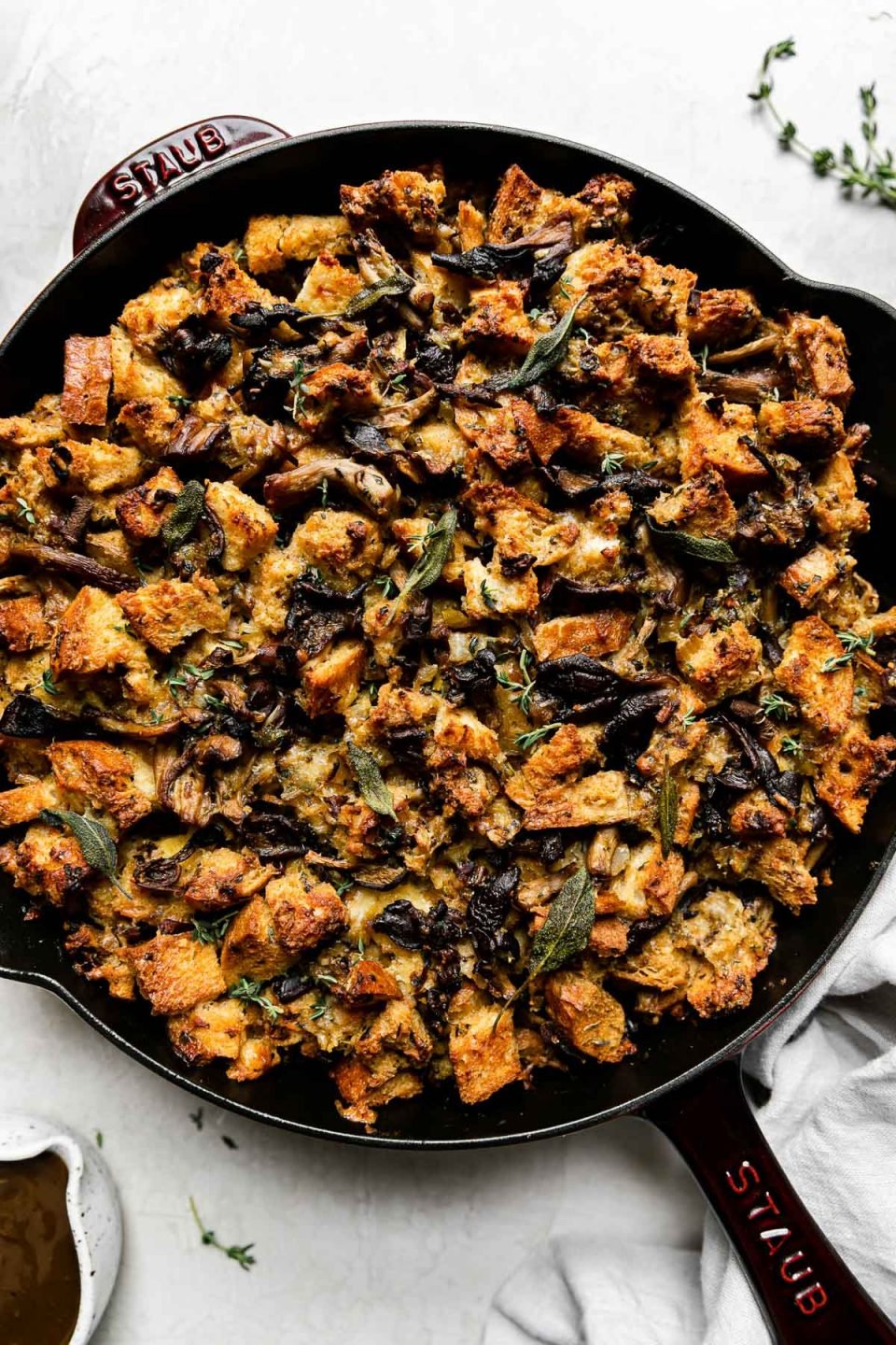 Baked wild mushroom stuffing fills a Grenadine colored Staub Cast Iron Skillet that sits atop a creamy white textured surface. A light gray linen napkin, loose sprigs of fresh herbs, and a small ceramic bowl filled with gravy sits alongside the skillet.