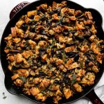 Baked wild mushroom stuffing fills a Grenadine colored Staub Cast Iron Skillet that sits atop a creamy white textured surface. A light gray linen napkin, loose raw wild mushrooms, sprigs of fresh herbs, and a small ceramic bowl filled with gravy sits alongside the skillet.