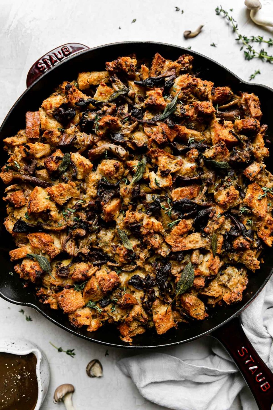 Baked wild mushroom stuffing fills a Grenadine colored Staub Cast Iron Skillet that sits atop a creamy white textured surface. A light gray linen napkin, loose raw wild mushrooms, sprigs of fresh herbs, and a small ceramic bowl filled with gravy sits alongside the skillet.