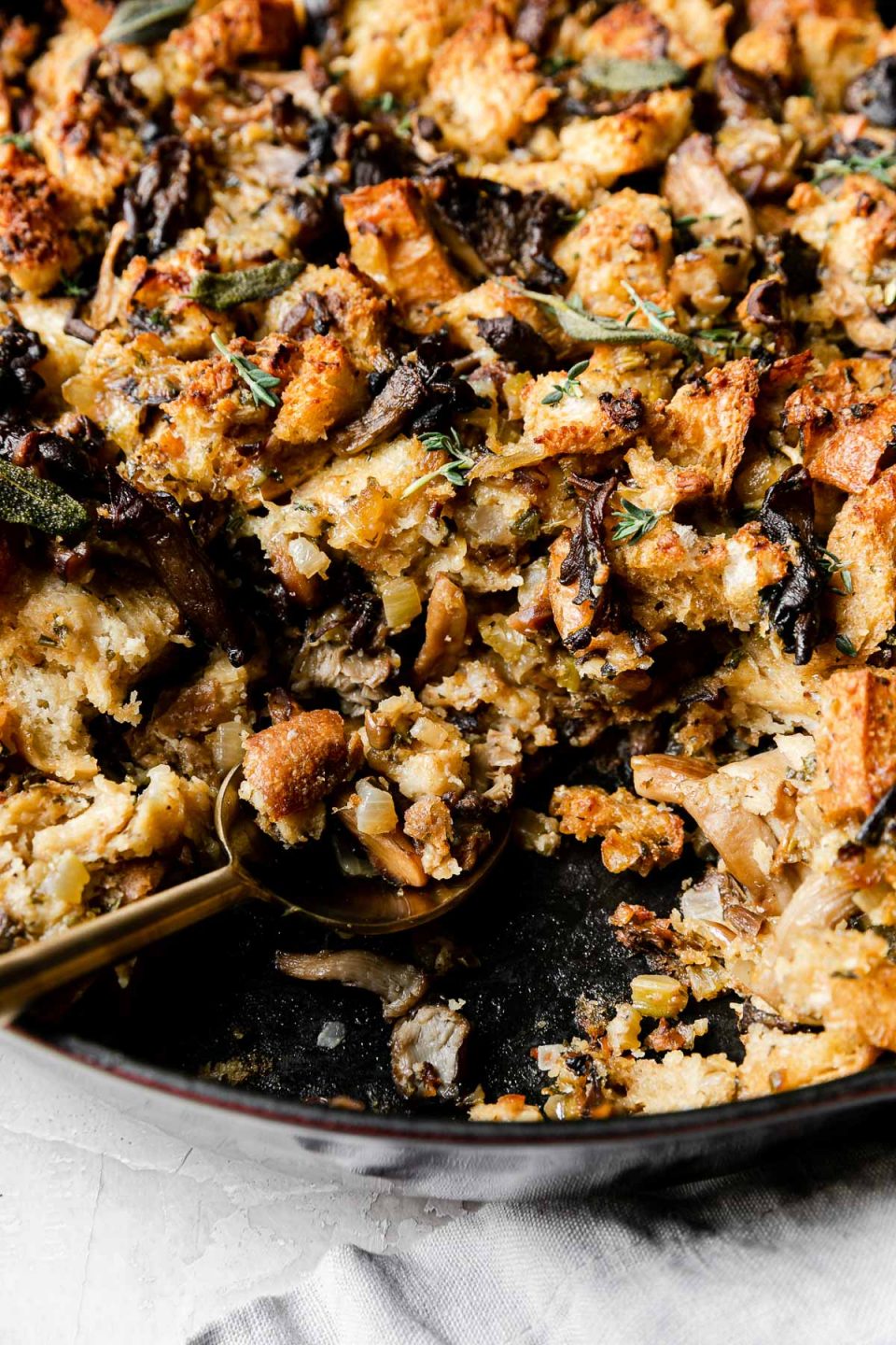 A side angle shot of Baked Crispy Wild Mushroom Stuffing with a portion scooped out fills a Grenadine colored Staub Cast Iron Skillet that sits atop a creamy white textured surface. A light gray linen napkin rests alongside the skillet and a gold serving spoon rests inside of the skillet.