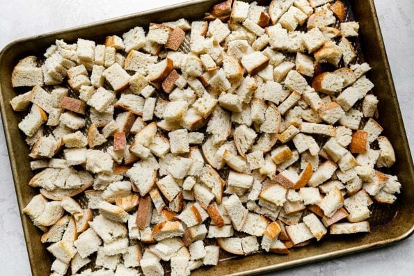 How to Make Crispy Cast Iron Skillet Wild Mushroom Stuffing, step 1: loaf bread cubes are cut and arranged on a baking sheet for toasting. The baking sheet sits atop a creamy white textured surface.