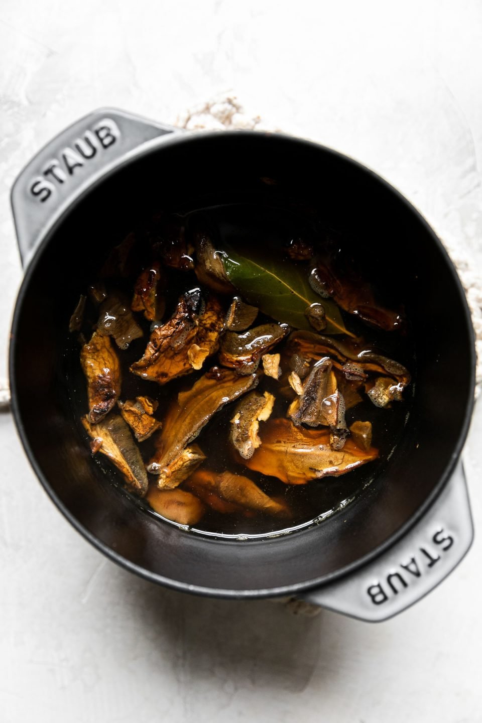 Dried porcini mushrooms reconstitute in vegetable broth with a bay leaf in a Staub Cast Iron Petite Dutch Oven. The small dutch oven rests atop a cream colored hot pad that sits atop a creamy white textured surface.