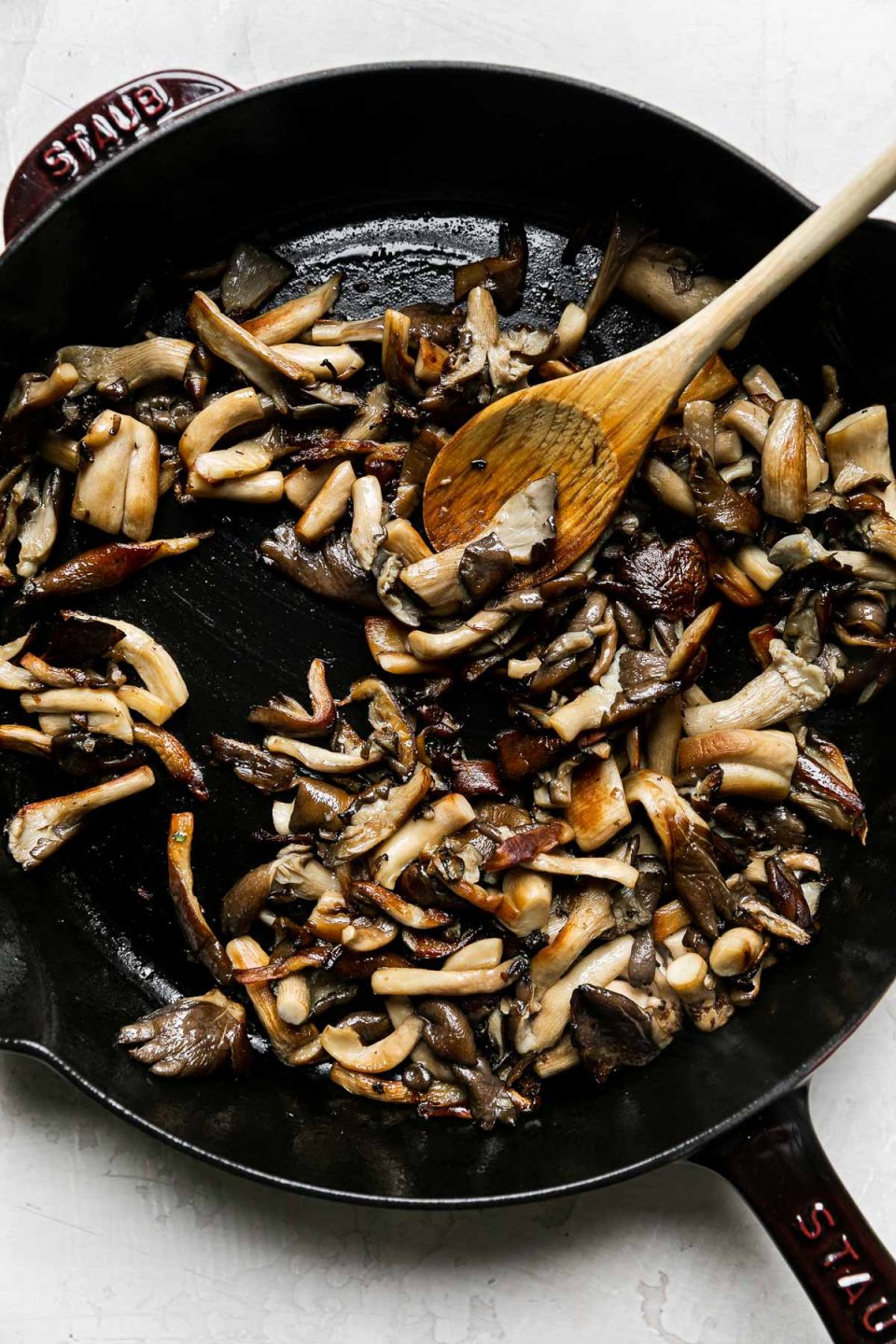 Sautéed & browned mushrooms in a Grenadine Staub Cast Iron Skillet. A wooden spoon rests in the skillet and the skillet sits atop a creamy white textured surface.