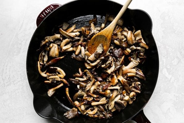 Sautéed & browned mushrooms in a Grenadine Staub Cast Iron Skillet. A wooden spoon rests in the skillet and the skillet sits atop a creamy white textured surface.