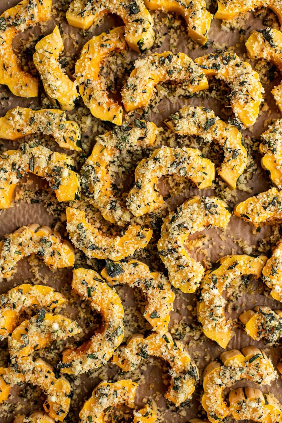 Slices of Delicata Squash seasoned with parmesan cheese, panko breadcrumbs, and fresh herbs are arranged on a piece of brown parchment paper for roasting.