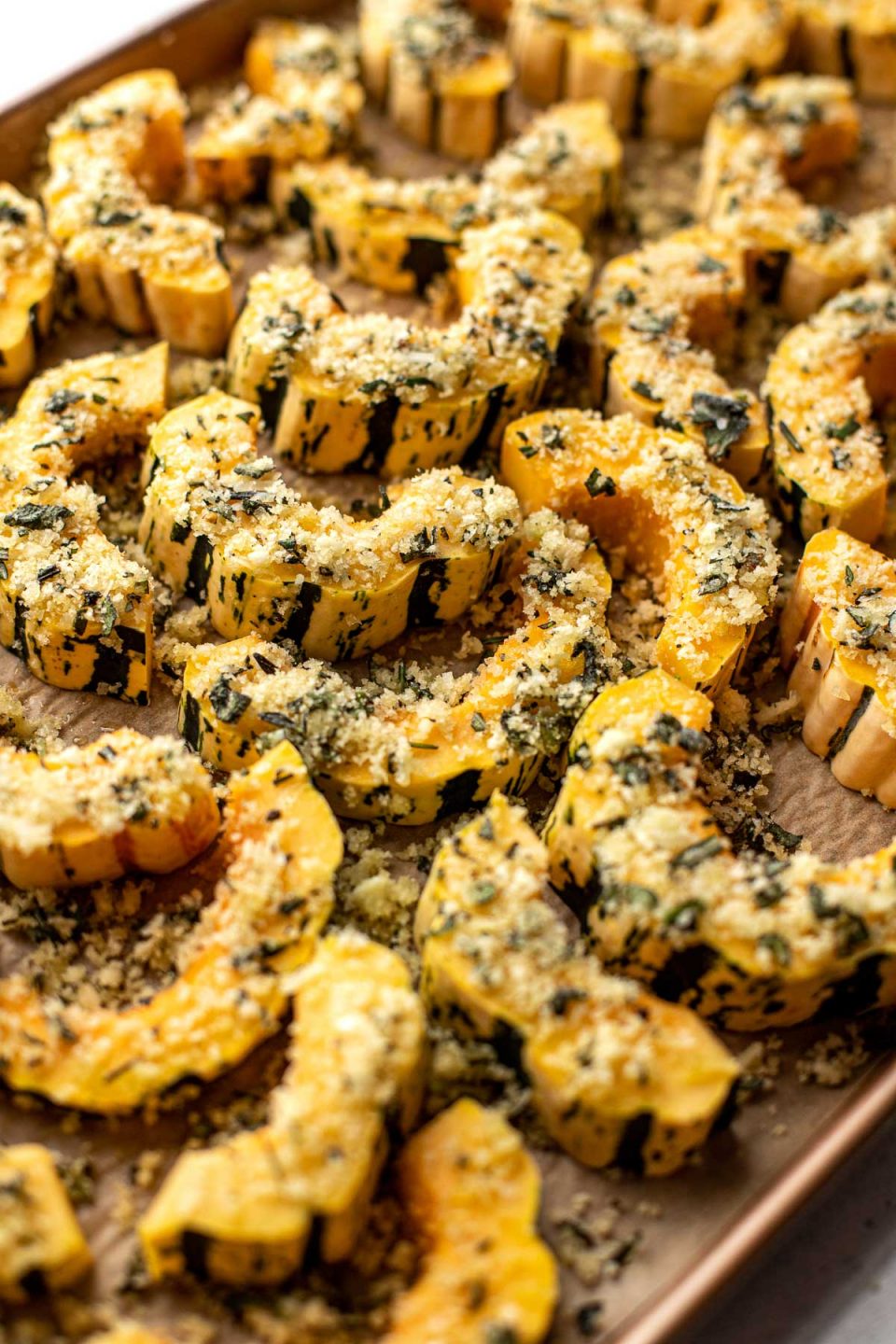 Slices of Delicata Squash seasoned with parmesan cheese, panko breadcrumbs, and fresh herbs are arranged on a baking sheet lined with brown parchment paper for roasting.